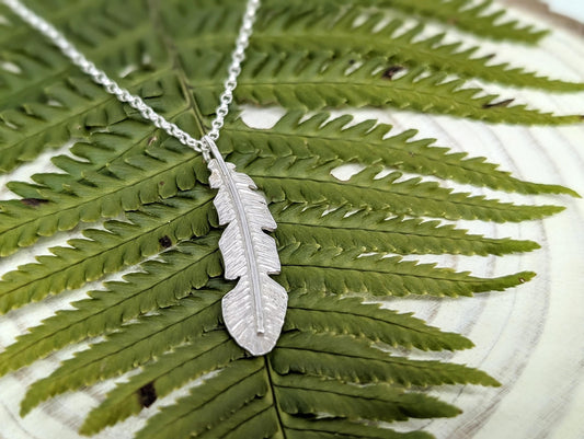 A sterling silver handmade feather pendant sits on top of a fern leaf.  The feather has a texture stamped into it and is hanging from a sterling silver belcher chain.