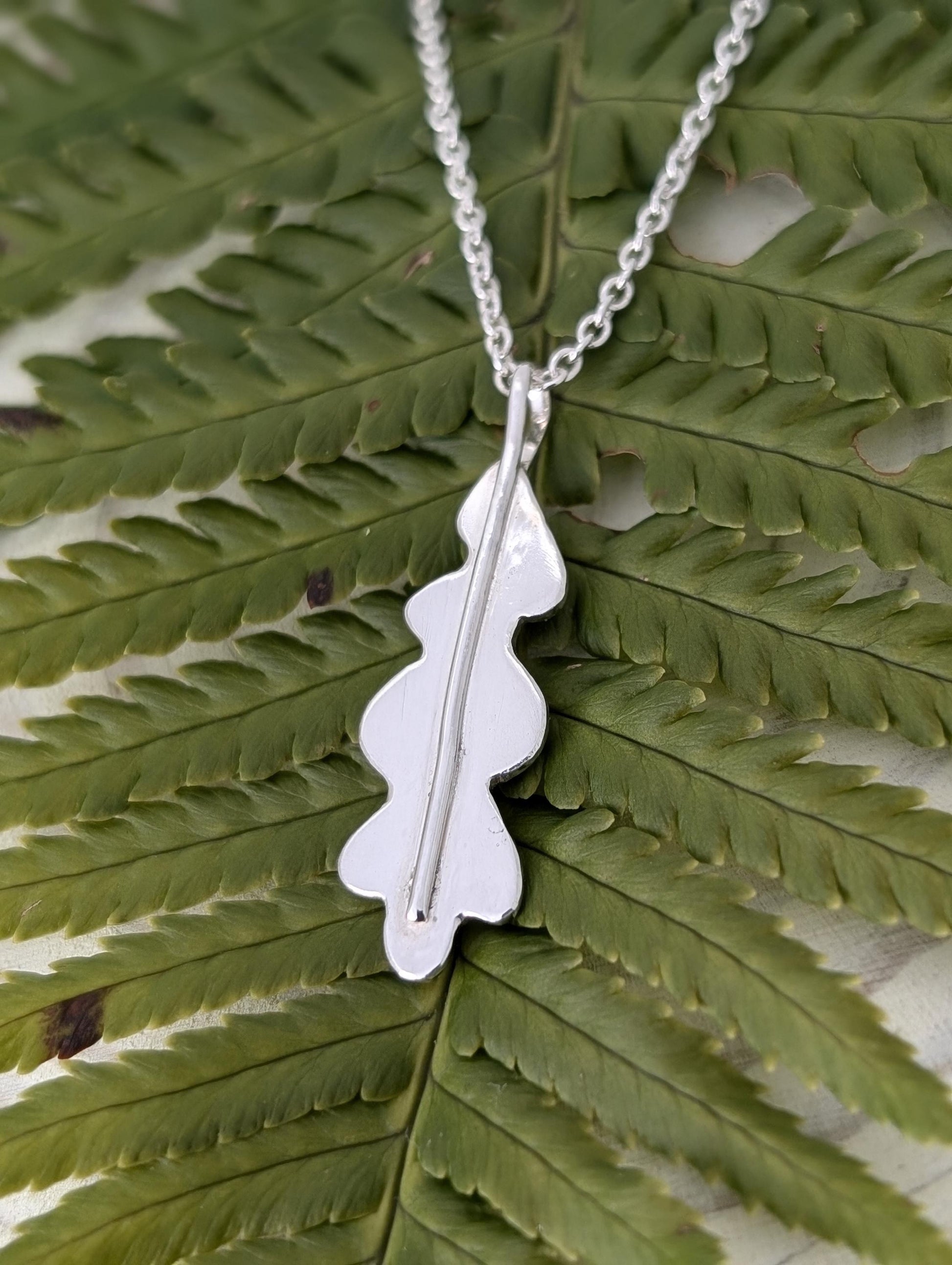 A handmade sterling silver oak leaf pendant hangs from a sterling silver belcher chain, resting on a fern leaf.