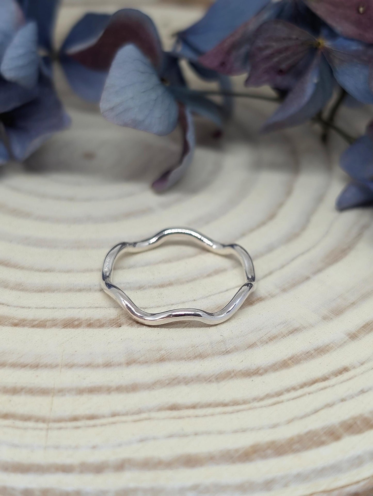 A silver ring shaped into a wavy pattern rests on a log slice with some blue hydrangeas in the background.