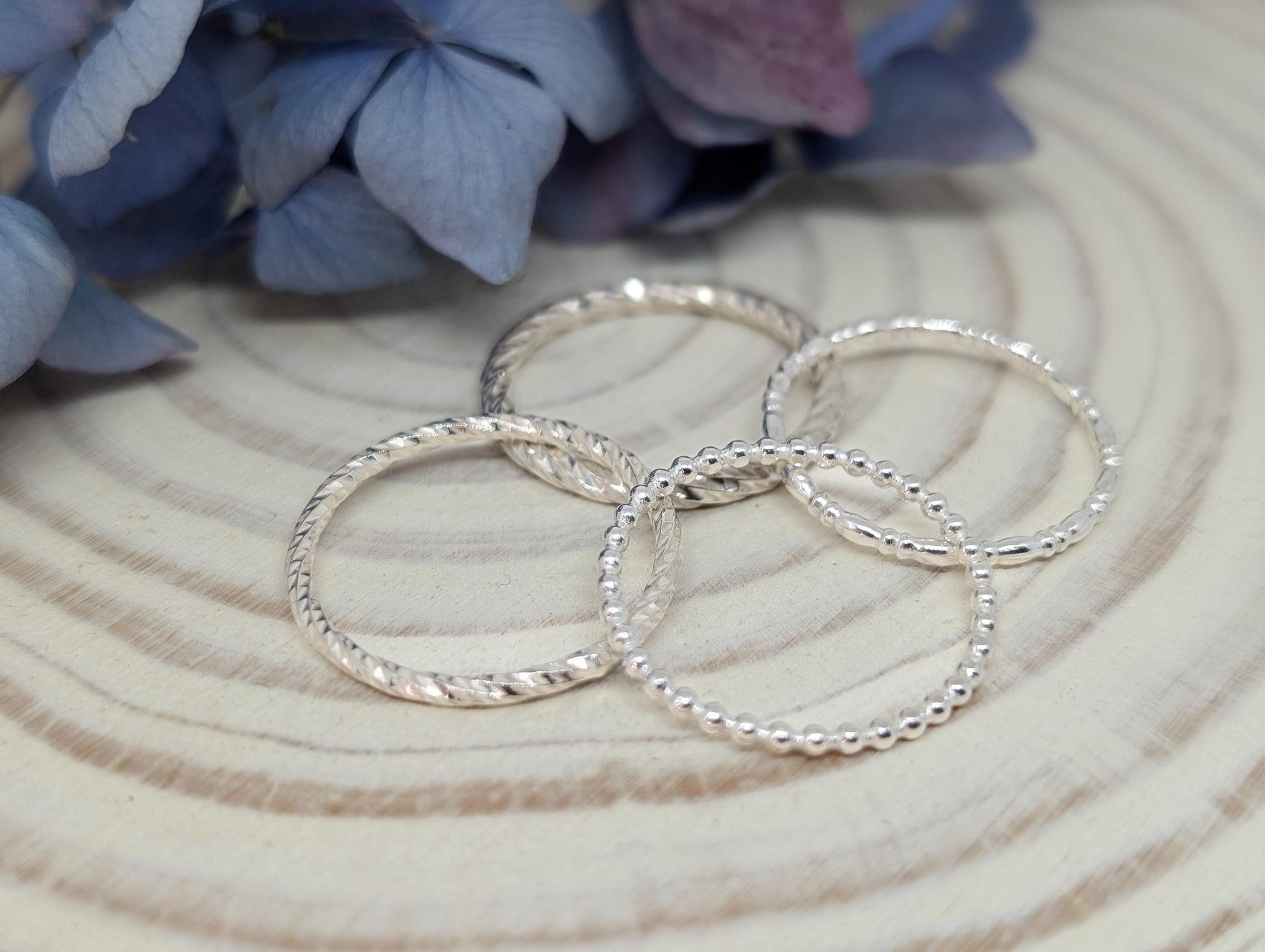 Four dainty silver rings rest on a log slice with some blue hydrangea flowers in the background. Each ring has a different pattern.