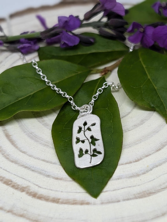 A sterling silver necklace featuring an intricate leafy branch cut out design. The pendant has a frosted surface and sits ontop of a green leaf.