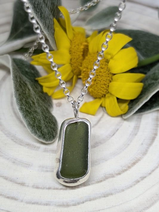 A small olive green piece of sea glass set in silver and attached to a silver chain to form a necklace.  It is displayed on a white log slice with yellow flowers and green leaves in the background.